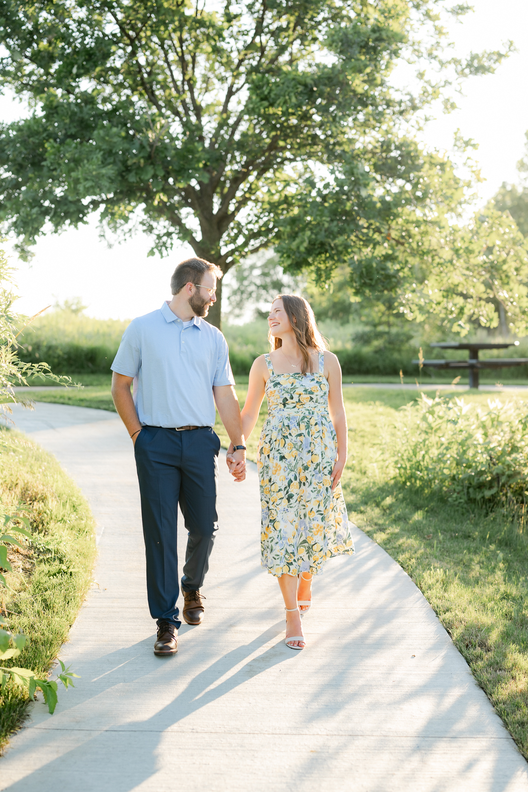 a man and woman holding hands and walking on a path for their summer engagement session in ames